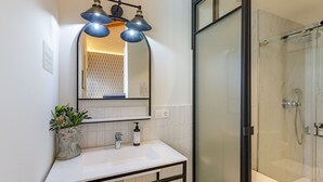 Een moderne badkamer met een witte wastafel, een spiegel met drie hanglampen, een douchecabine met glazen wanden en een potplant op de wastafel.