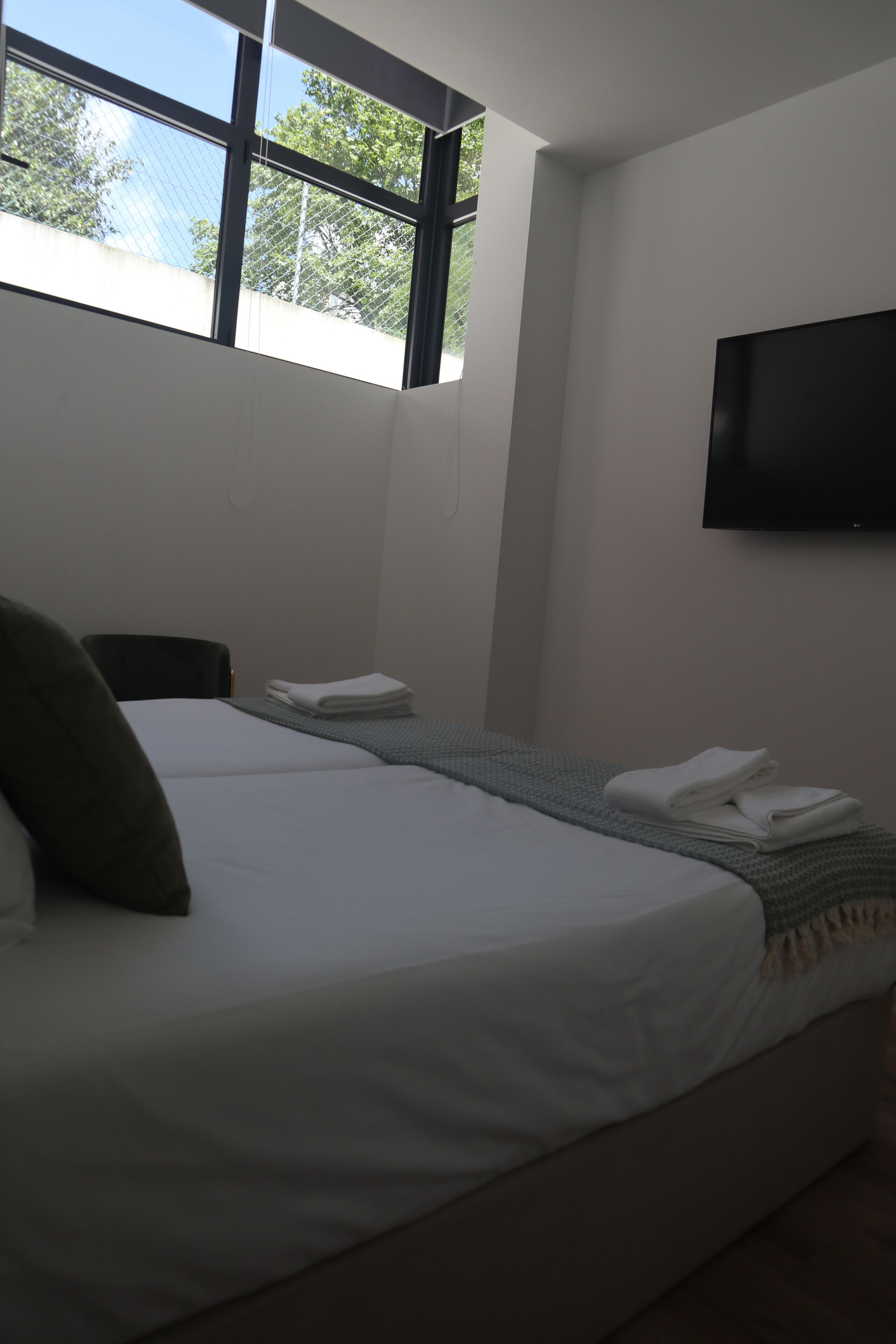 A hotel room with a bed, a chair, a flat-screen TV mounted on the wall, and a window with a view of trees.