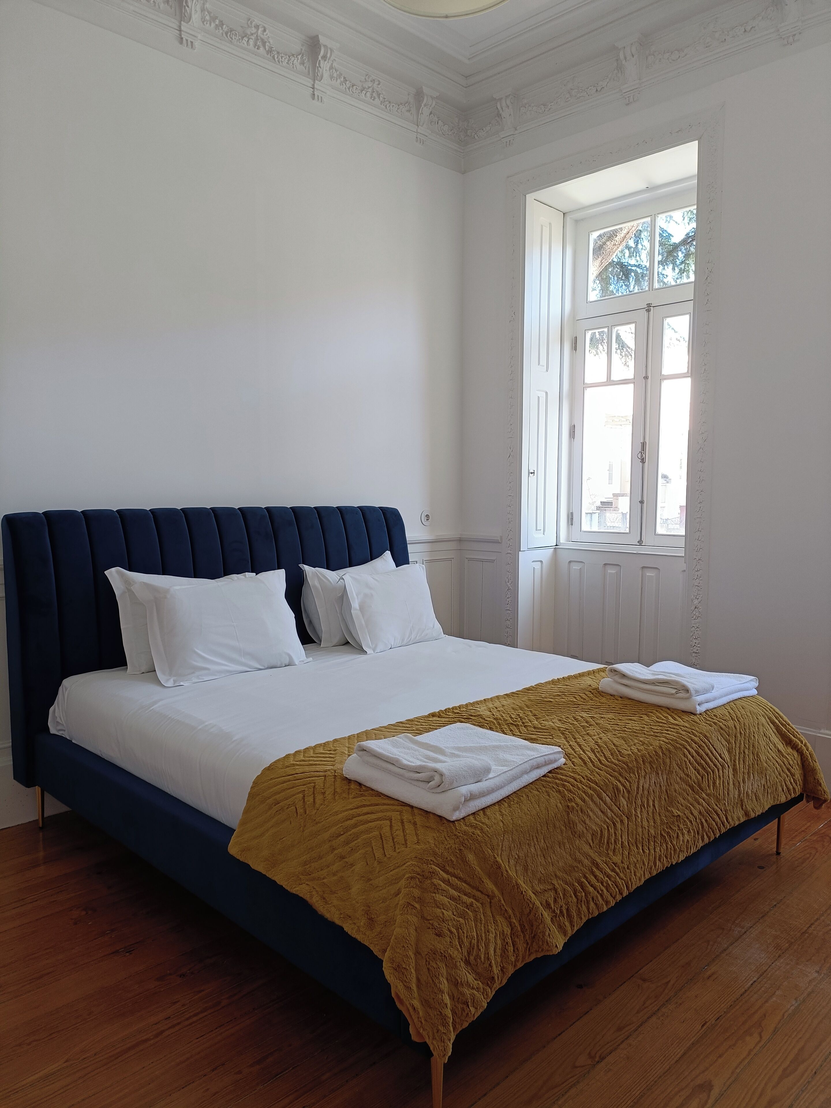 A neatly made bed with white linens and a mustard-colored blanket, a dark blue headboard, and a window with white shutters.