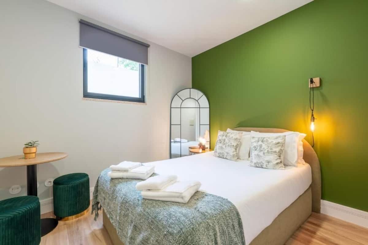 A bedroom with a bed, a small round table with a plant, a green ottoman, a window with a blind, and a wall-mounted light fixture.