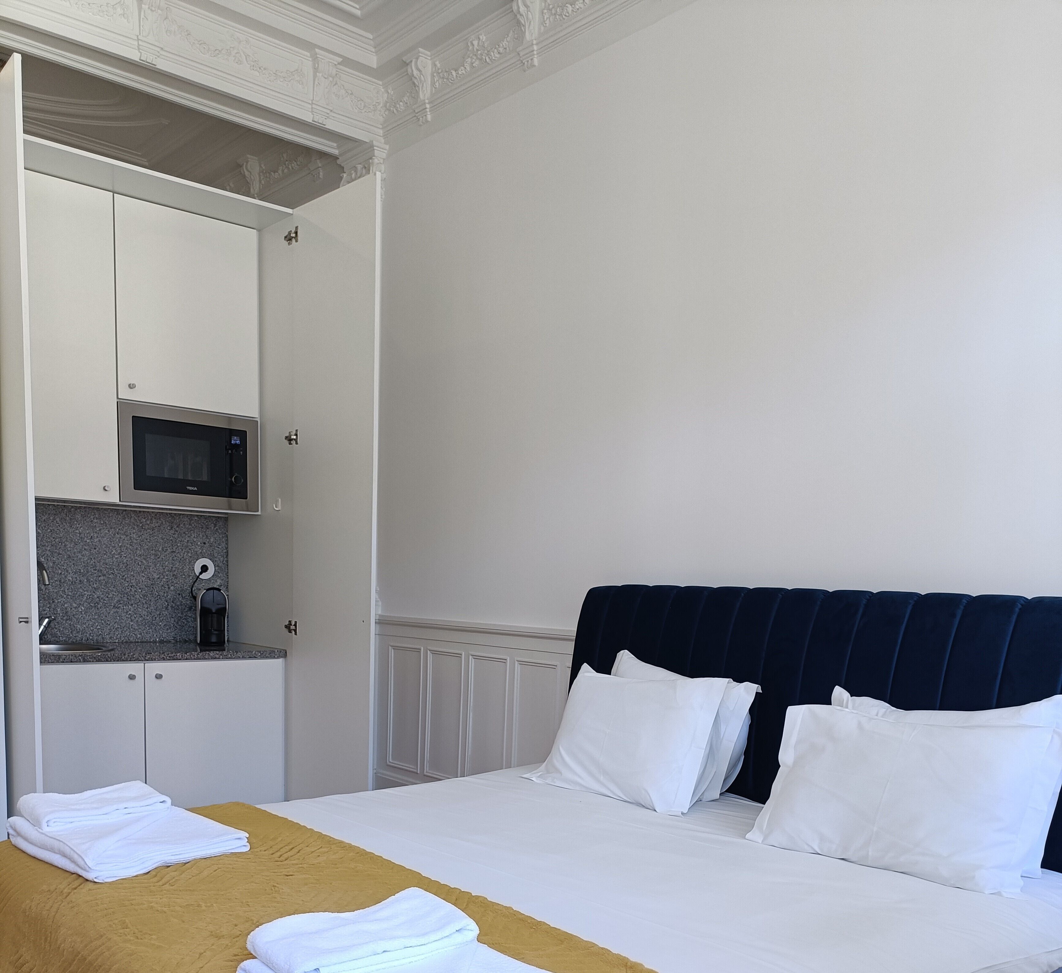 Habitación de hotel con cama, mesita de noche con toallas, microondas, pequeña cocina y cabecero azul oscuro.
