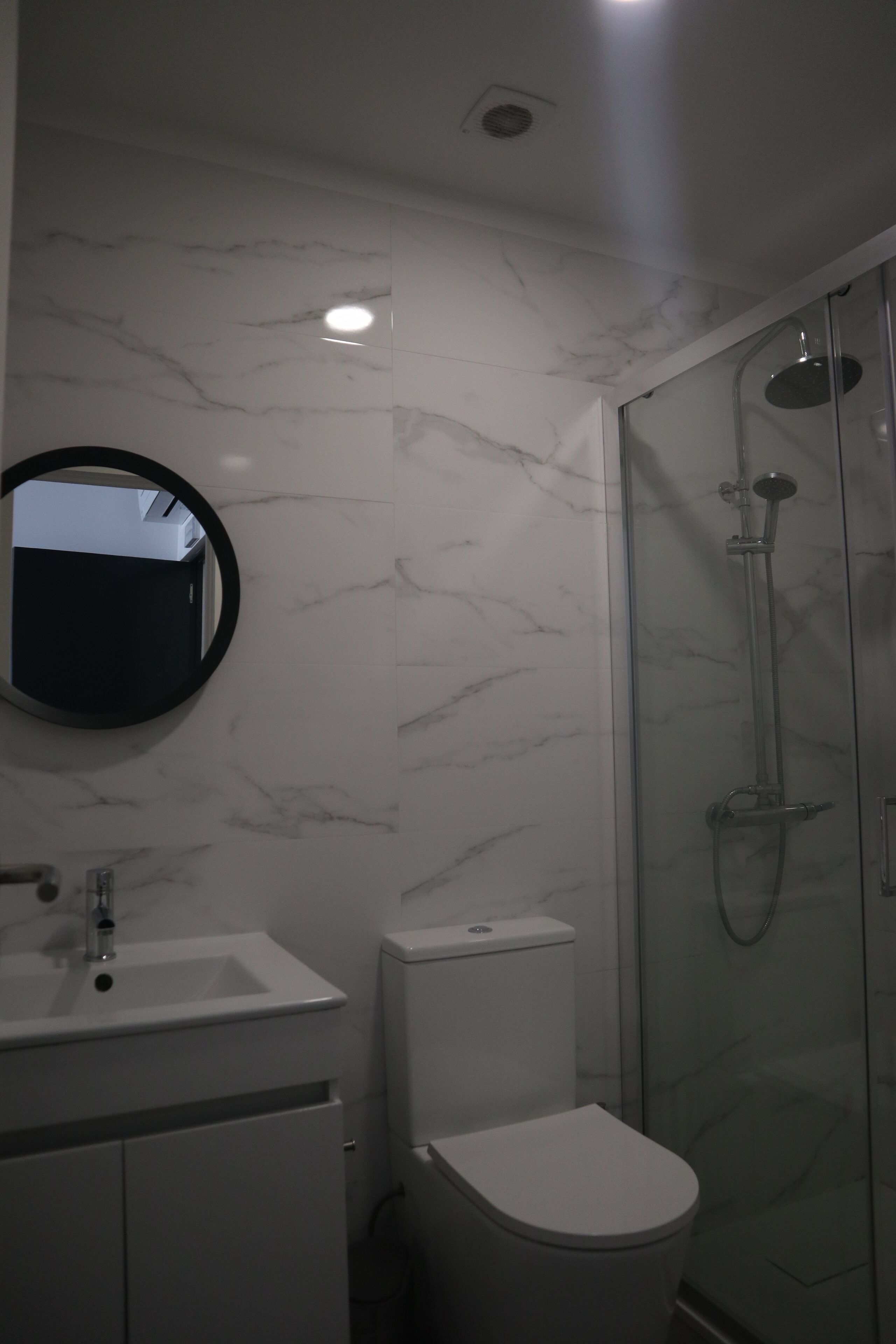 A bathroom with a marble wall, a round mirror, a sink, a toilet, and a glass-enclosed shower area.