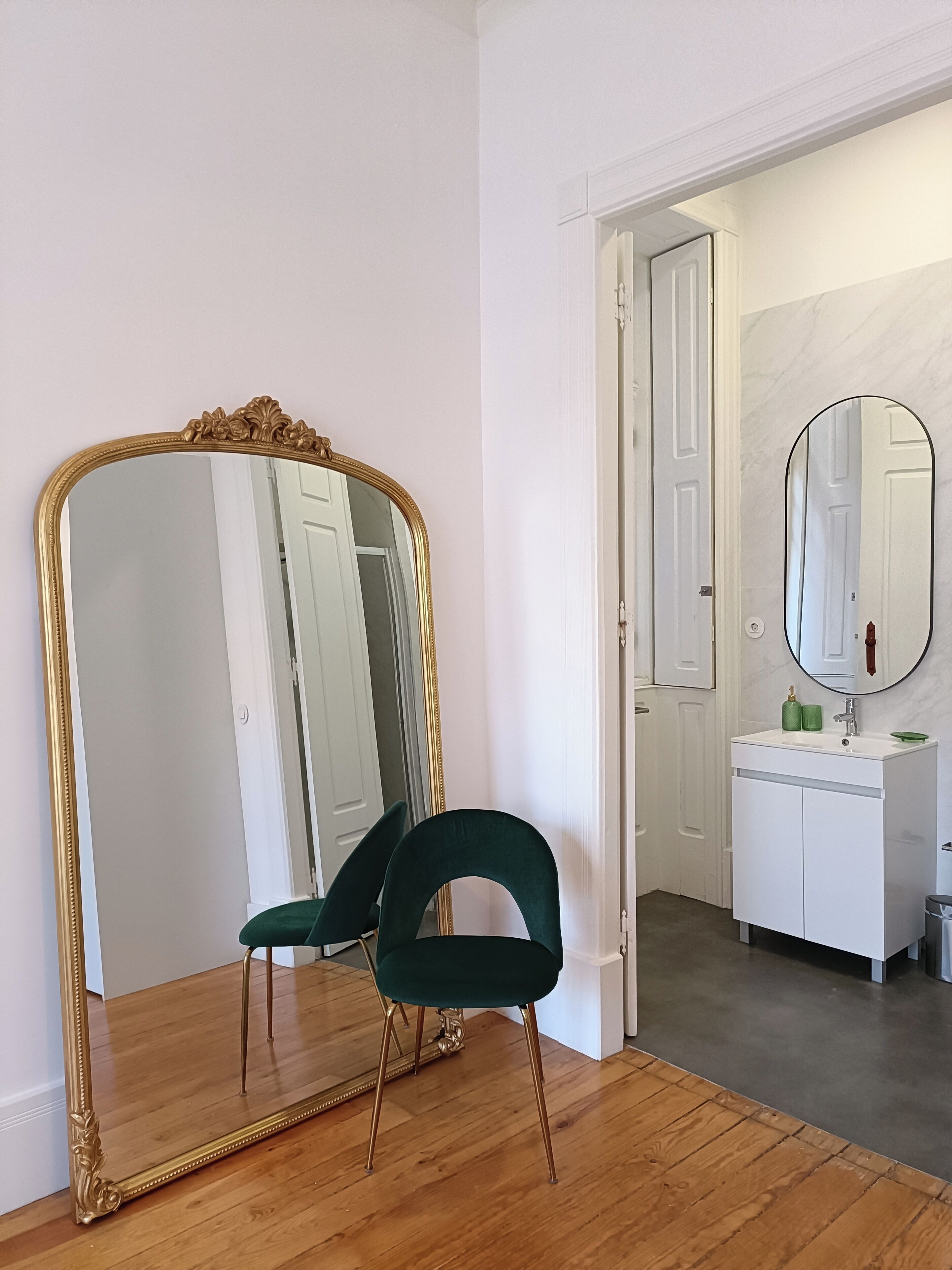 A room with a large ornate mirror, a green upholstered chair, a white dresser, and a bathroom visible through an open door.
