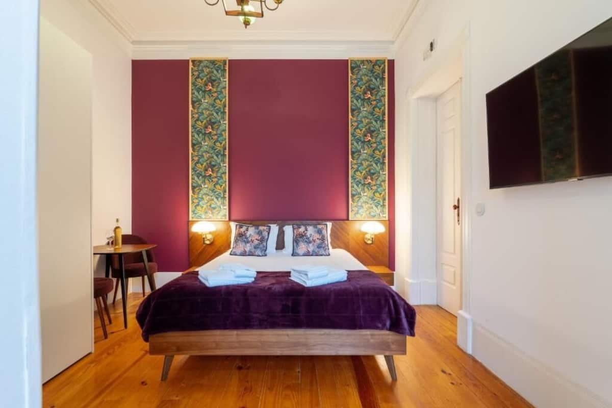 A bedroom with a wooden bed, two bedside lamps, a TV on the wall, and a small table with chairs.