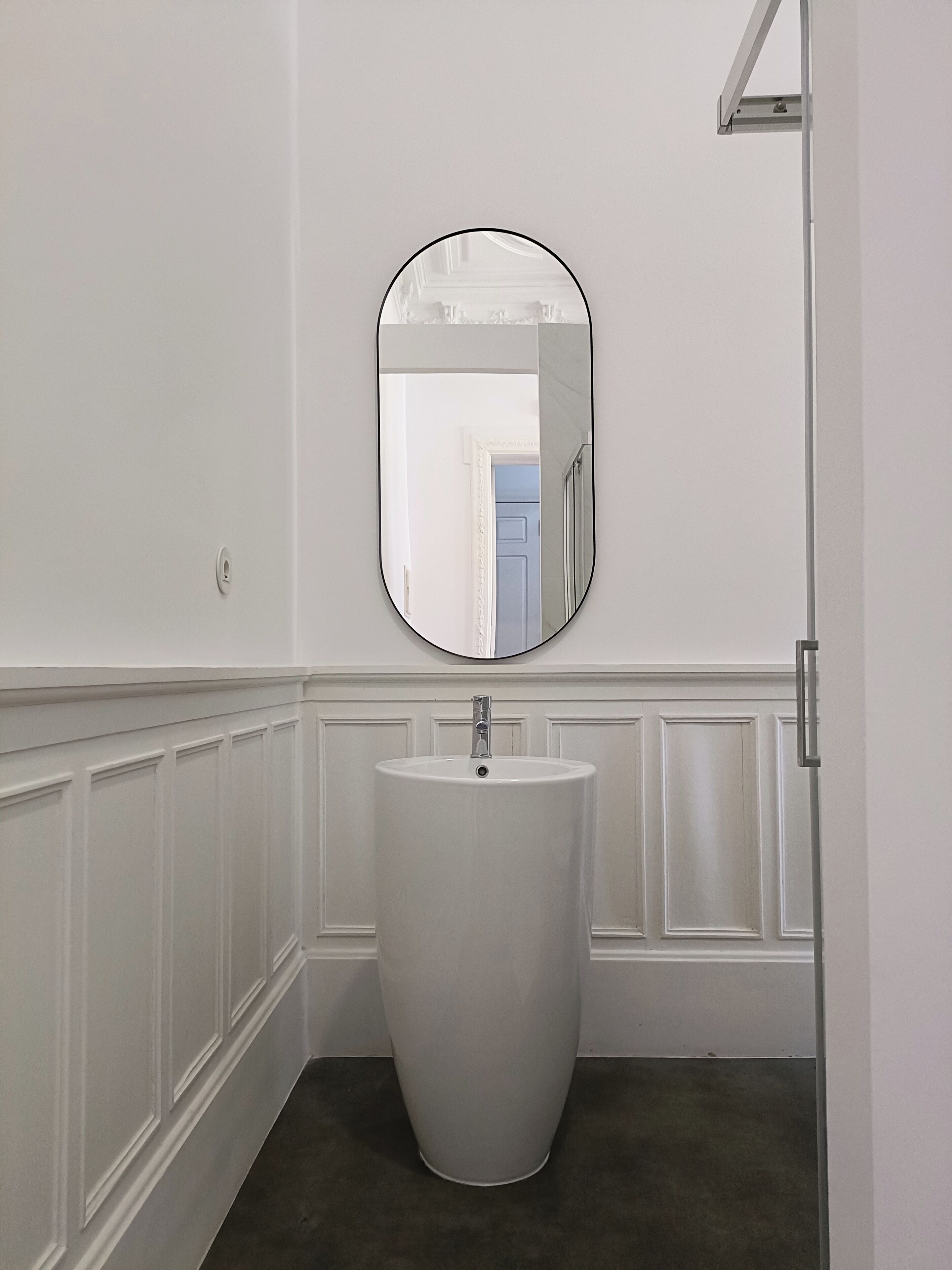 A bathroom with a pedestal sink, a wall-mounted oval mirror, and a door leading to another room.