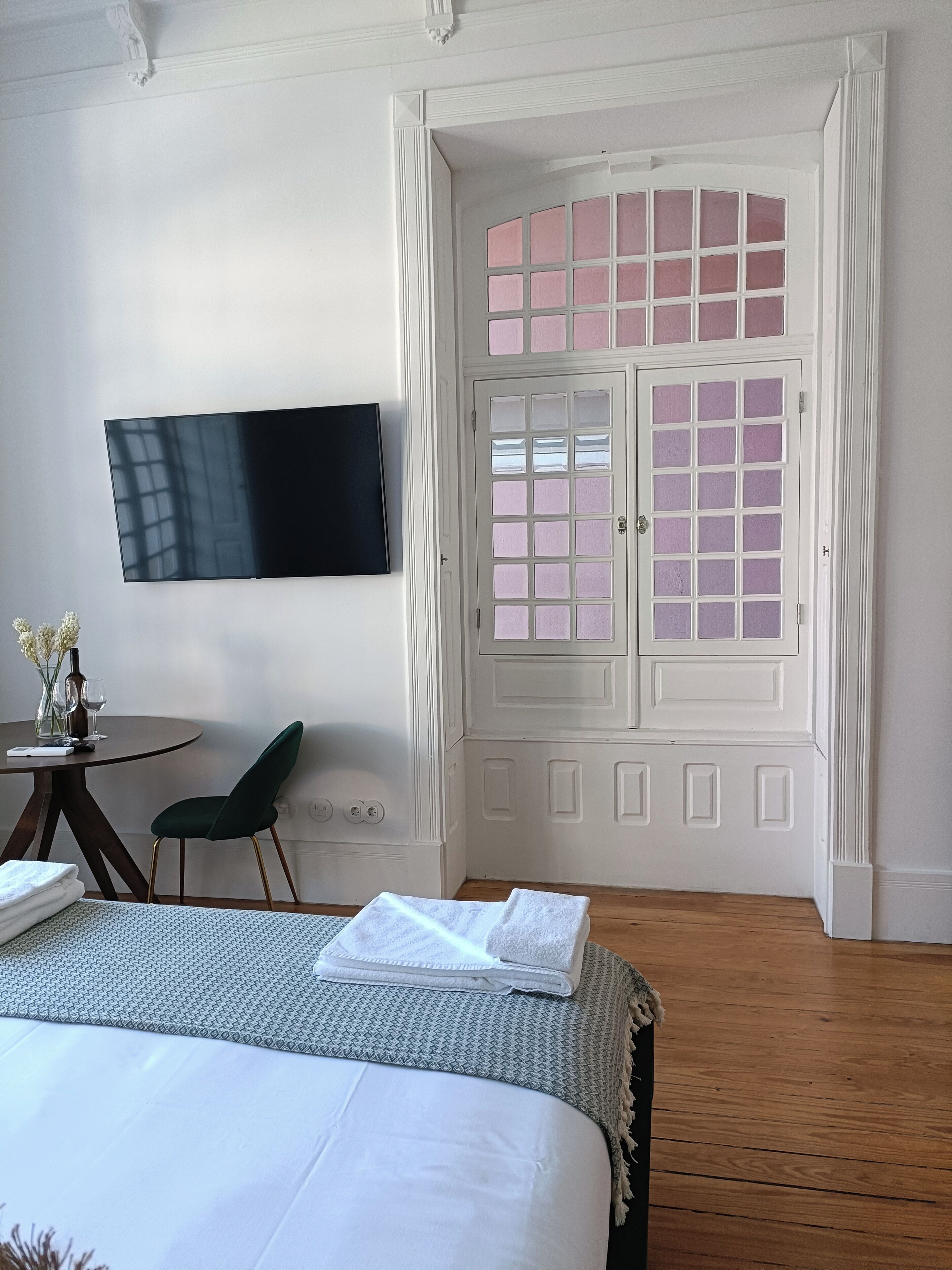 Une chambre d’hôtel avec un lit, un téléviseur fixé au mur, une petite table avec une chaise et une fenêtre avec une porte blanche.