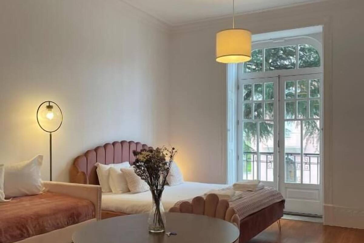 A modern bedroom with a bed, a round table with chairs, a floor lamp, and a window with a view of greenery.