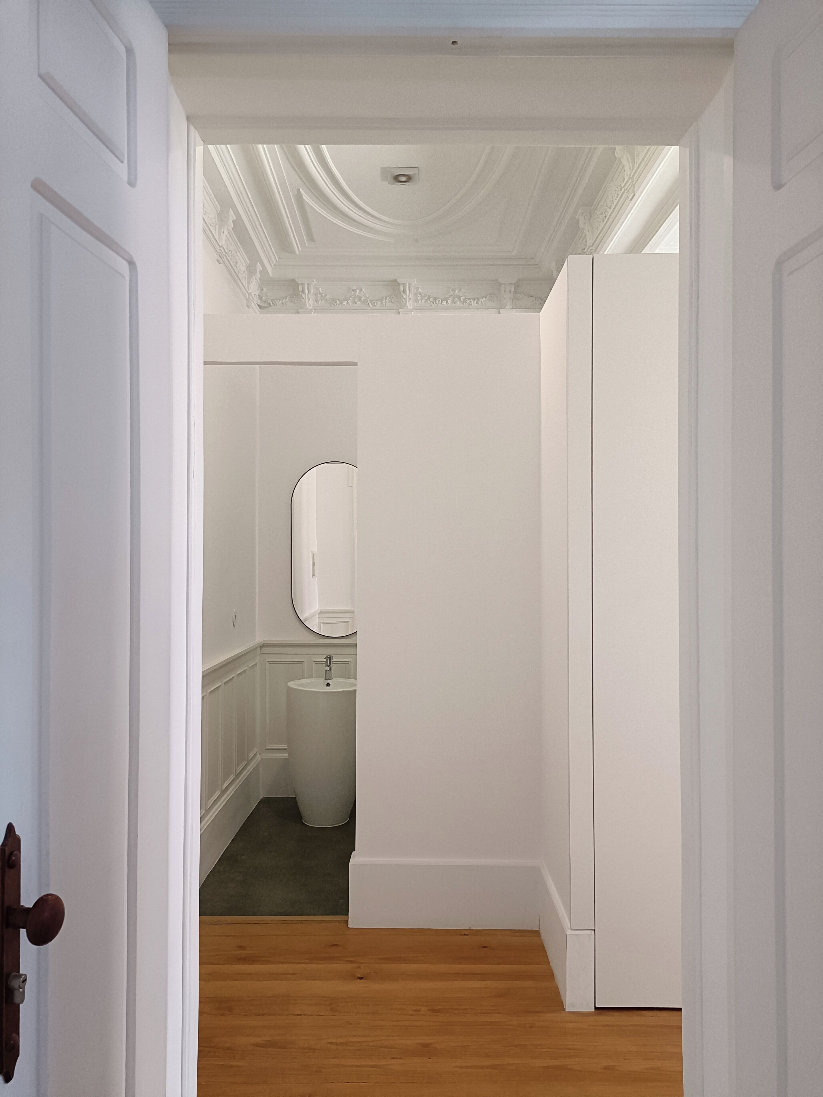 A narrow hallway with a wooden floor leading to a bathroom with a white sink and a mirror.