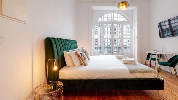 Un dormitorio moderno con un cabecero tapizado en verde, una cama colgante, una mesita redonda pequeña y un ventanal con vistas a edificios.