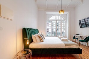 Un dormitorio moderno con un cabecero tapizado en verde, una cama colgante, una mesita redonda pequeña y un ventanal con vistas a edificios.