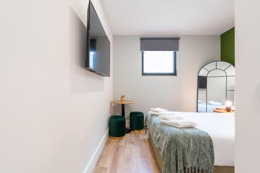 A bedroom with a bed, a small round table, two green ottomans, a wall-mounted TV, a window, and a mirror.