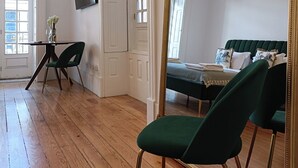 Una habitación con piso de madera, una silla tapizada de verde, una cama con cabecera verde, un espejo grande y ornamentado, y un televisor en la pared.