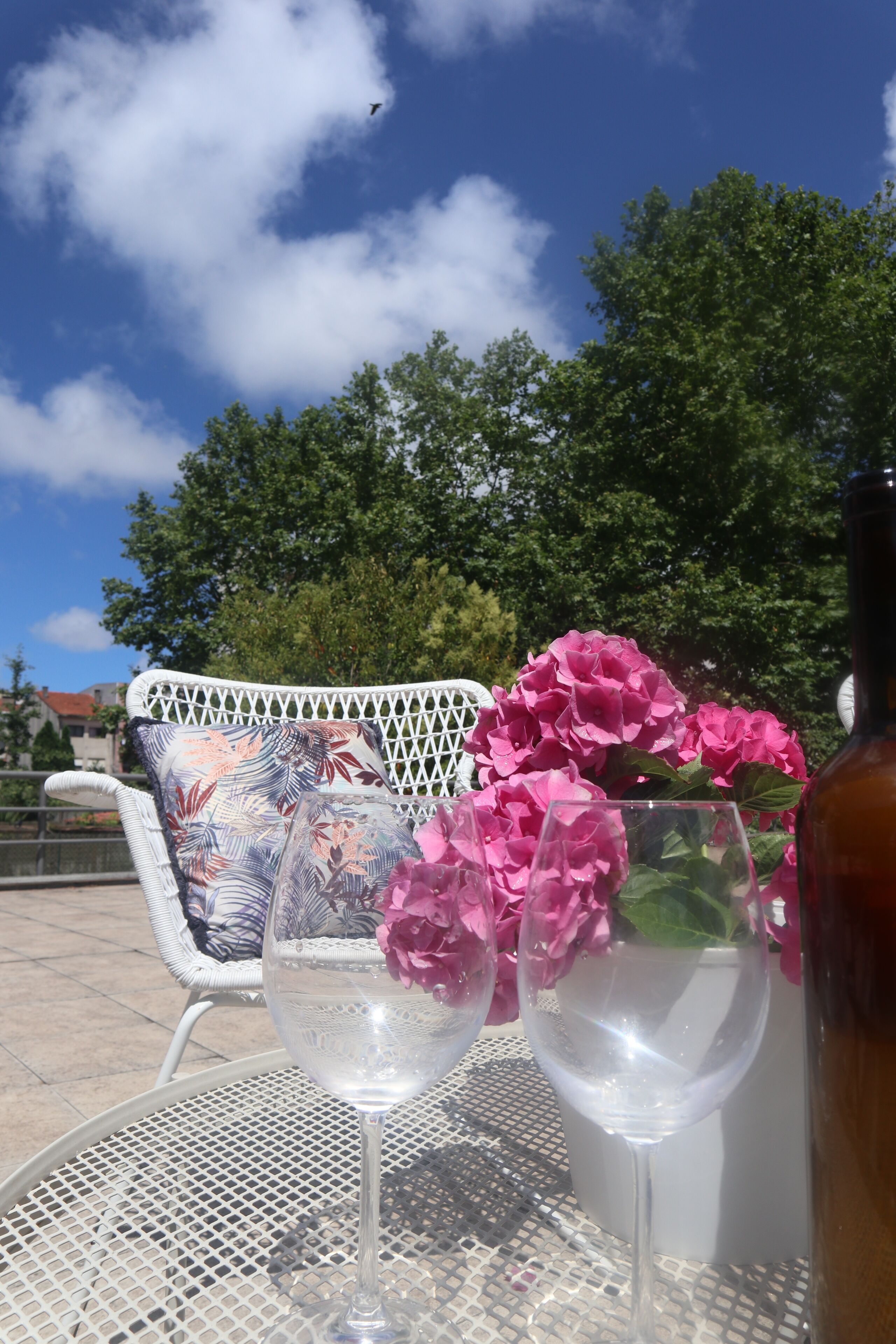 Ein Tisch mit zwei Weingläsern und einer Flasche, umgeben von rosa Blüten, draußen mit einem weißen Stuhl und Grünpflanzen im Hintergrund.