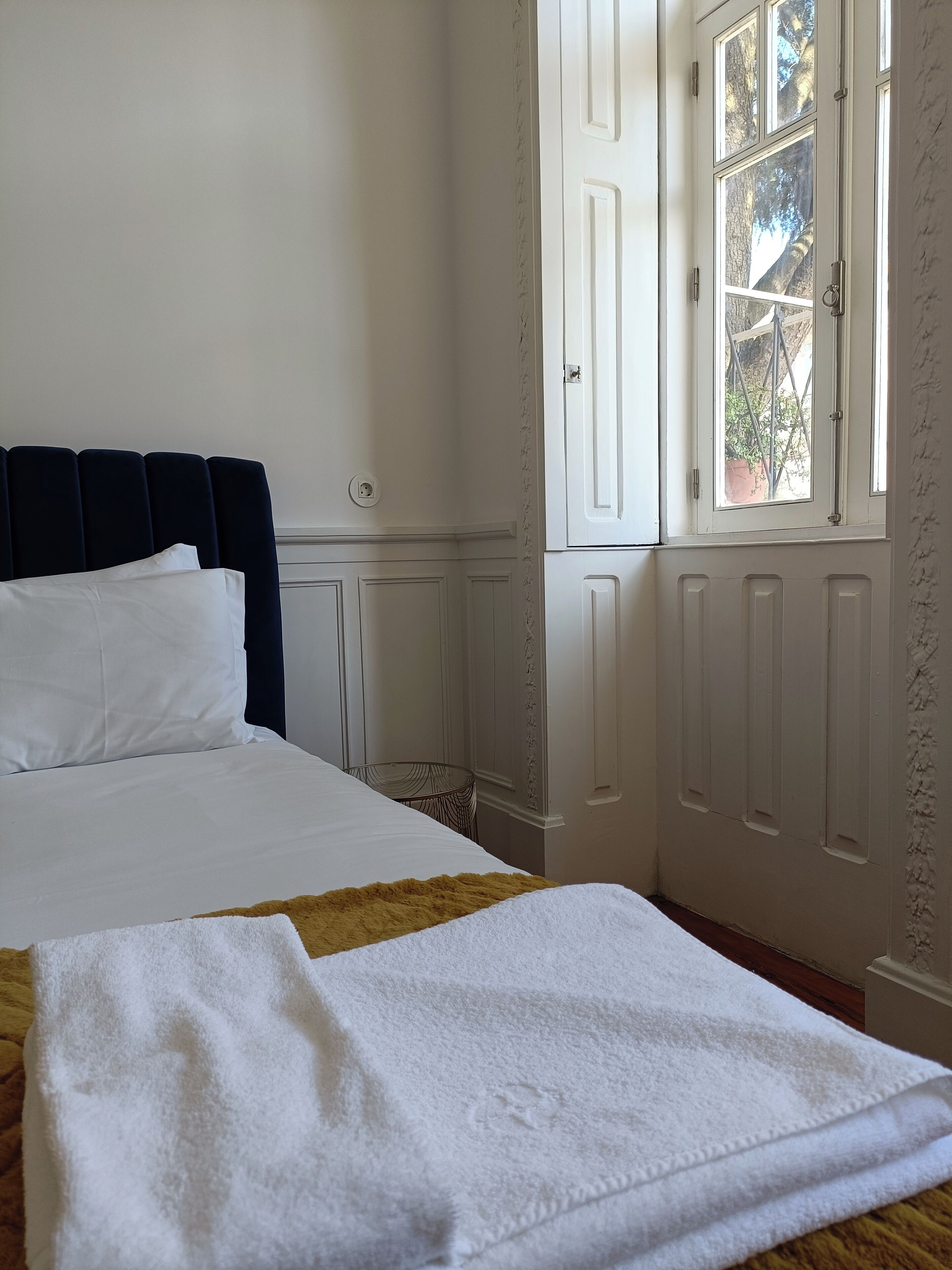 A neatly made bed with white linens, a dark headboard, and a window with a view of greenery outside.