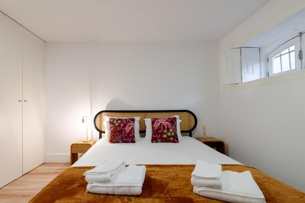 Un dormitorio con cama, dos almohadas, una mesita de noche, una lámpara y una ventana.