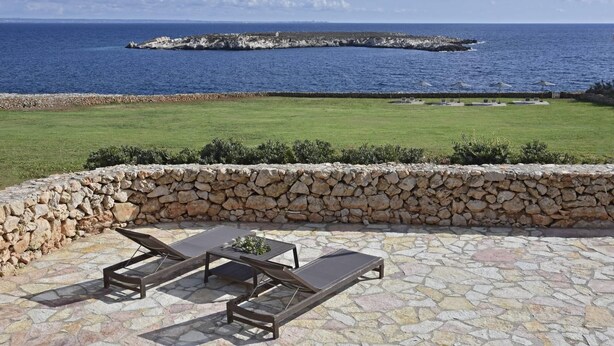 A stone patio with a table and lounge chairs overlooking a grassy area and the sea.