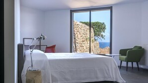 A modern bedroom with a large bed, a desk, and a view of a rocky coastline.
