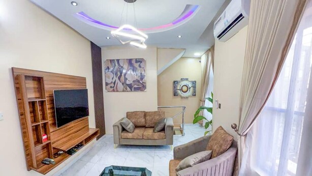 A modern living room with a flat-screen TV, a sofa, a coffee table, and a unique glass water feature. - Home Away From Your Home (Lagos)