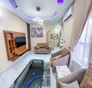 A modern living room with a flat-screen TV, a sofa, a coffee table, and a unique glass water feature. - Home Away From Your Home (Lagos)