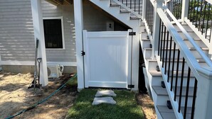 A two-story house with a white exterior, a front porch, and a staircase with black railings. - Cute, cozy and newly built bungalow on private street awaits your Capecod visit. (Mashpee)