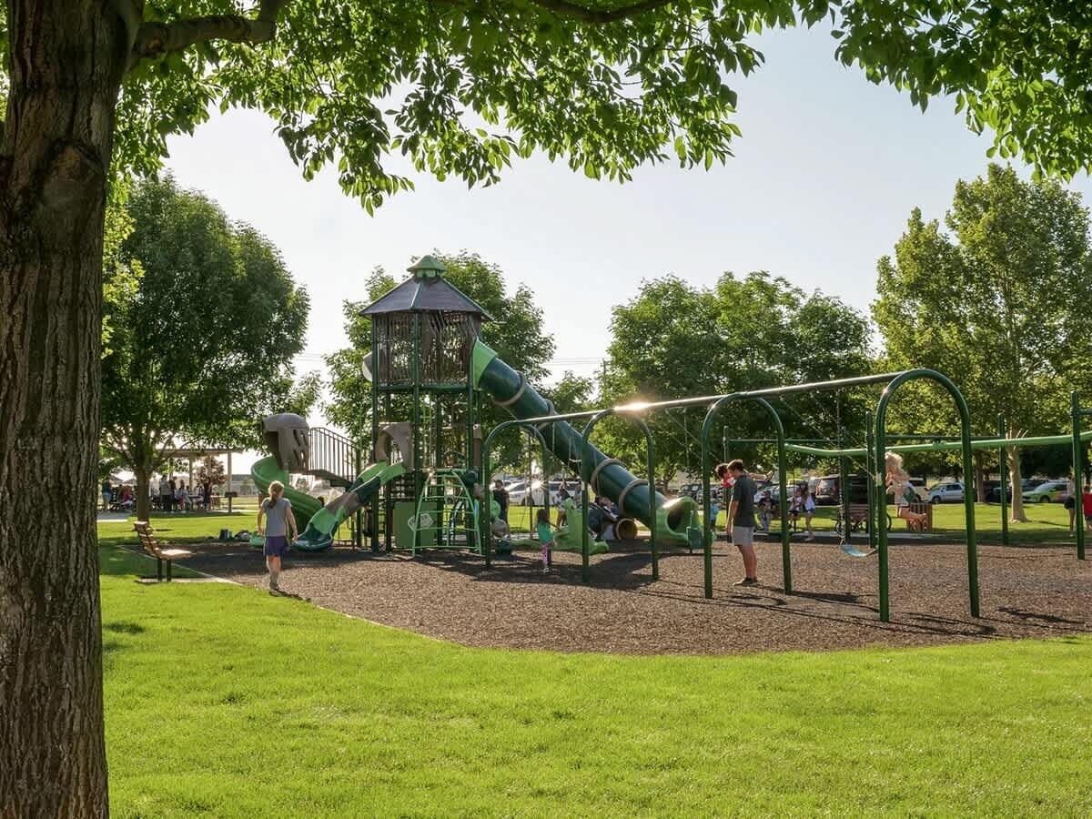 Une aire de jeux avec des toboggans verts et des balançoires, entourée d’arbres et comprenant un espace pique-nique.