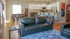 A modern living room with a black leather sofa, a dining table with chairs, and a kitchen area with white cabinets and stainless steel appliances. - Sleeps 25 | Theater Room | Near Roaring Springs | Large Meridian Home (Meridian)