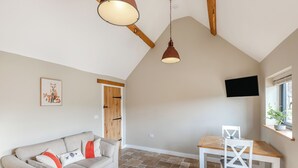 A loft-style room with a high ceiling, exposed wooden beams, a sofa, a dining table, and a TV mounted on the wall.