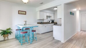 A modern kitchen with white cabinets, a blue island, and a dining area with a blue table and red stools. - Walk to Pier: St Augustine Beach Condo w/ Balcony! (St. Augustine Beach)