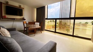 A living room with a gray sofa, a dining table with chairs, a flat-screen TV mounted on the wall, and a large window with a view of palm trees and a beach. - Flat FM20 Cabo Branco Lotus - By Almare (João Pessoa)