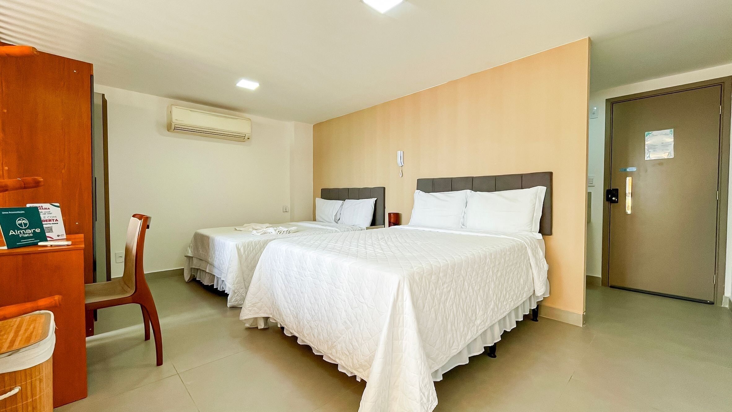 A hotel room with two beds, a wooden door, a chair, an air conditioning unit, and a phone on the wall.