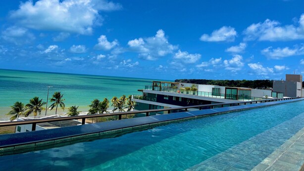 A modern hotel with a long swimming pool, a beach view, and a clear blue sky. - Flat FM20 Cabo Branco Lotus - By Almare (João Pessoa)