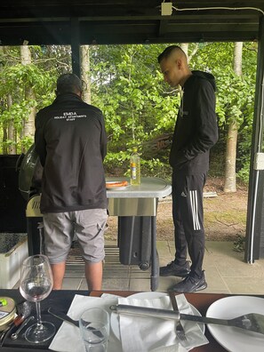 Two individuals at an outdoor dining area with a table set for a meal, surrounded by greenery.