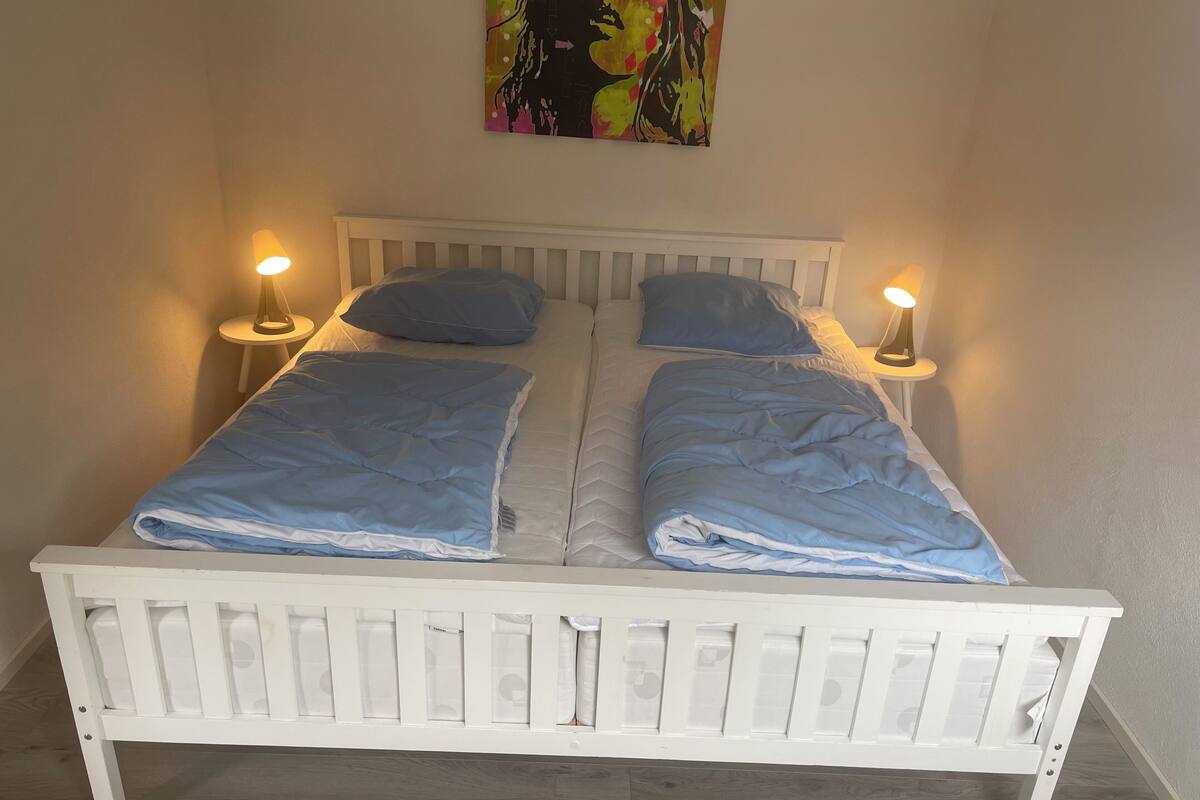Uma cama de casal com cabeceira e peseira brancas, dois travesseiros e duas luminárias de cabeceira.