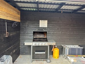 A portable grill with a gas tank and a metal container, set against a dark wooden wall with a metal roof.
