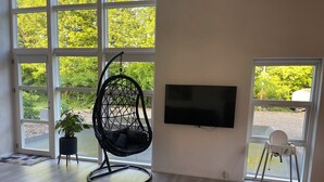 A modern living room with a large window, a black hanging chair, a flat-screen TV, and a small table with a chair. - EMDA country living close to Legoland. (Billund)