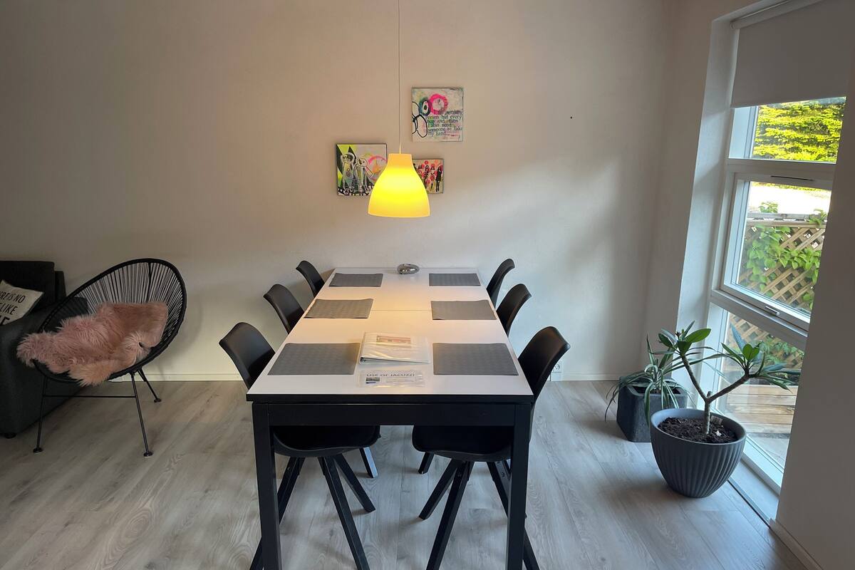 Uma sala de jantar com mesa posta para seis, cadeiras pretas, uma cadeira rosa e um abajur. Há plantas em vasos e obras de arte nas paredes.