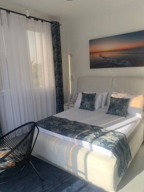 Un dormitorio con cama, una silla, una mesita de noche, un cuadro y una ventana con cortinas.