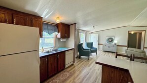 A compact kitchen with wooden cabinets, a refrigerator, and a sink. There is a dining area with a table and chairs, and a small seating area with a chair. - Home in Surf City- Pelicans Perch (Hampstead)