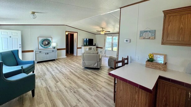 A spacious living room with a wooden floor, a blue armchair, a white sofa, a dining table with chairs, and a kitchen area with wooden cabinets and a countertop. - Home in Surf City- Pelicans Perch (Hampstead)