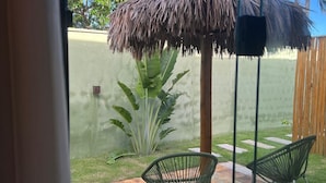 A view through a window of a patio area with a thatched umbrella, two green chairs, and a banana plant. - Villas Gran (Paraipaba)