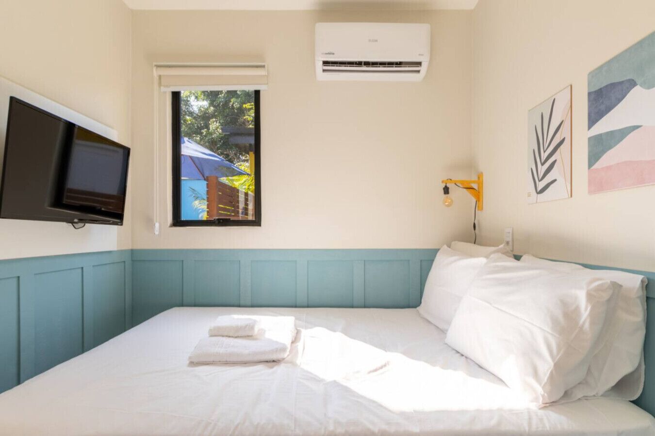 A bedroom with a bed, a television, an air conditioner, and a window with a view of a building and trees.