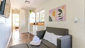 Una sala moderna con un sofá gris, una mesita con toallas dobladas, una zona de cocina con fregadero y armarios, y arte abstracto en la pared.