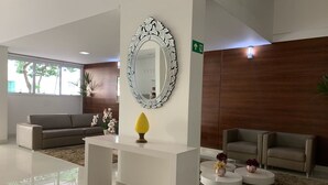 Een moderne lobby met een wit receptiebalie, een grote sier spiegel, en een grijze bank.
