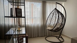 Een woonkamer met een hangstoel, een glazen plank met ingekaderde foto’s en een raam met dunne gordijnen.