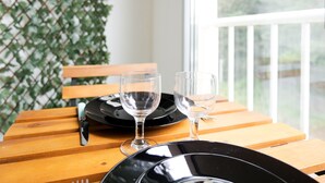 A wooden table with black plates and clear wine glasses, set against a backdrop of a white railing and greenery.