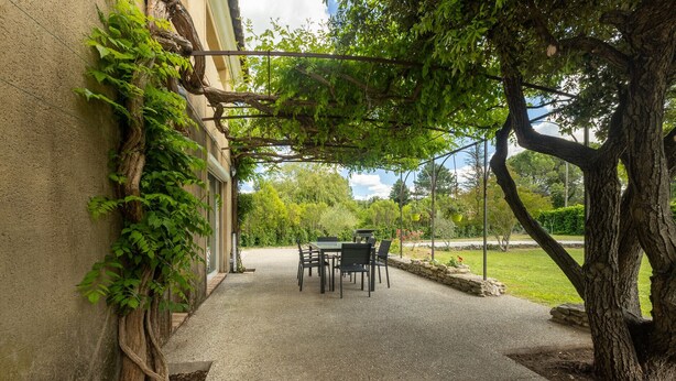 Eine überdachte Terrasse mit Tisch und Stühlen, umgeben von Grünpflanzen und einem Gebäude.