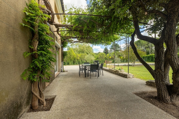 A covered patio with a table and chairs, surrounded by greenery and a building.