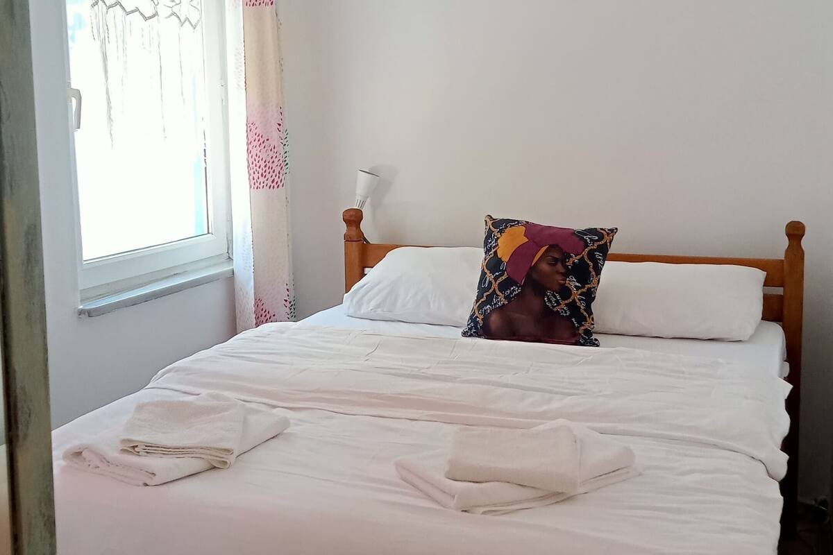 A bedroom with a bed, white bedding, a window with patterned curtains, an air conditioning unit, and a bedside lamp.