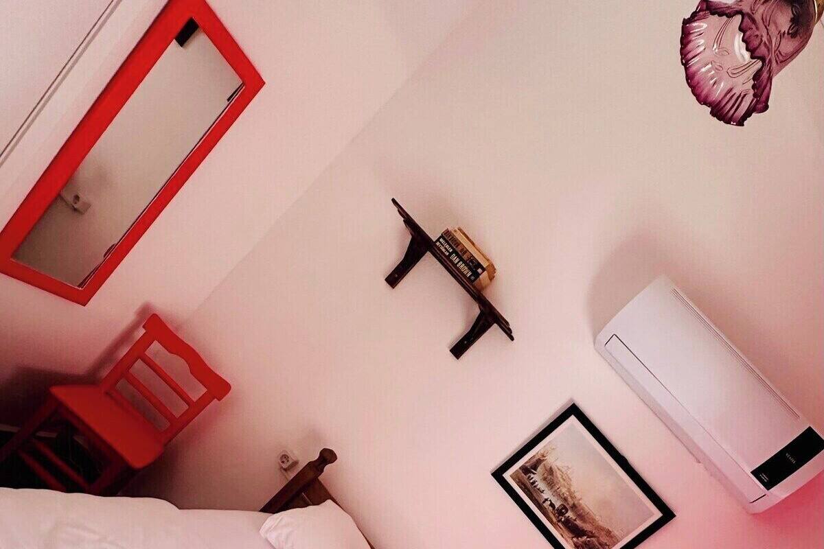 A bedroom with a bed, a red chair, a mirror, a picture frame, and an air conditioning unit.