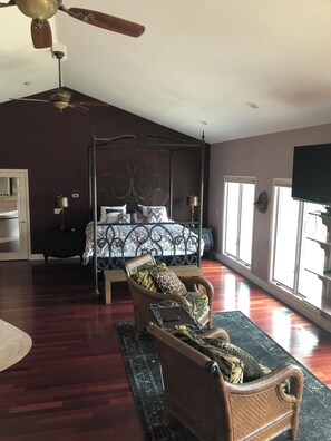 A spacious bedroom with a four-poster bed, a wicker chair, a ceiling fan, and large windows.